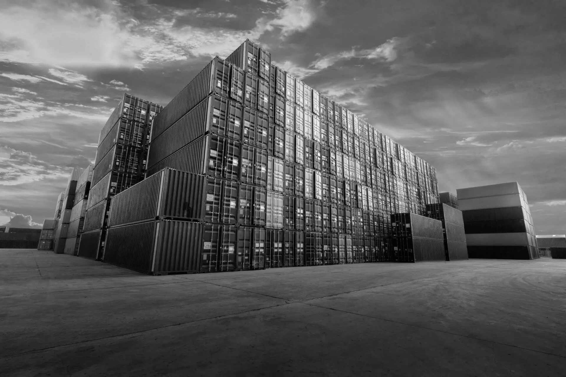 Vue en noir et blanc d'un immense mur de conteneurs maritimes empilés dans un terminal portuaire, sous un ciel nuageux